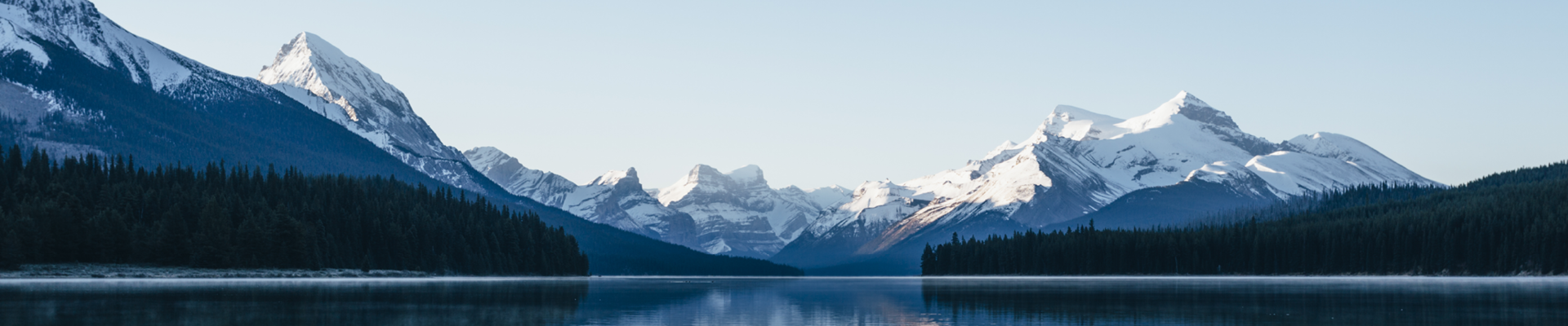 Large dark pond with dark green trees in the distance and snowy mountains in the back 
